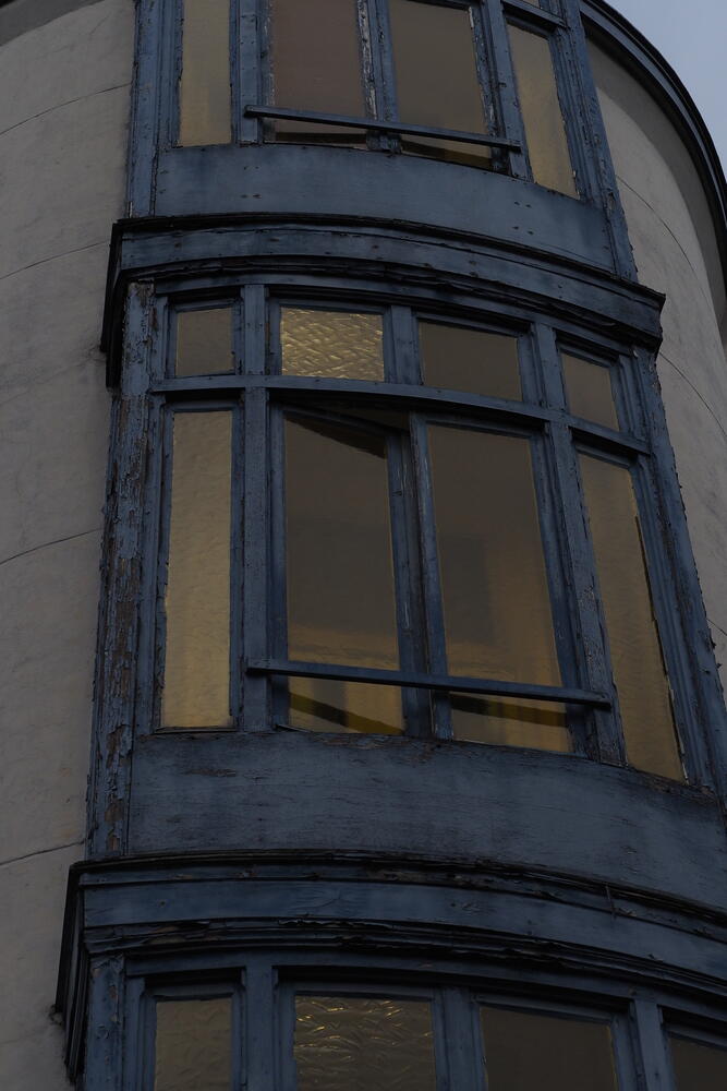 A small section of the side of a house showing its central, blue-framed windows. The paint is old and peeling off. The glasses appear to have a slightly yellow tint.